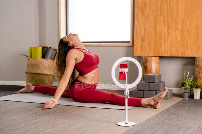 Side view of flexible barefooted woman in stylish red top and leggings performing Front Splits while recording vlog on smartphone attached to tripod in modern studio — Stock Photo