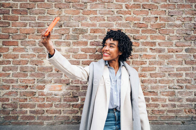 Mujer afroamericana positiva con ropa casual y abrigo sonriendo felizmente mientras toma autorretrato en el teléfono móvil - foto de stock