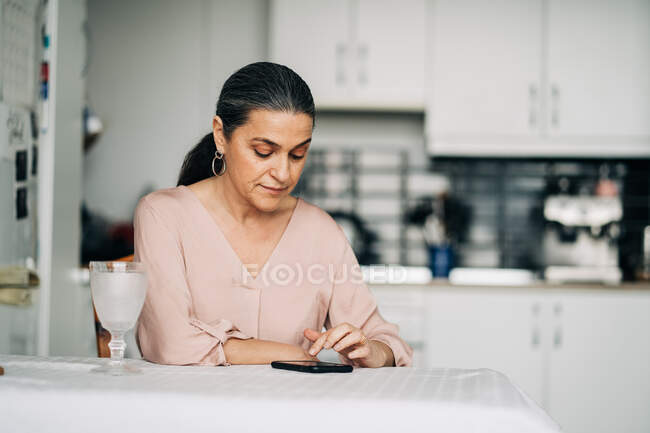 Cellulaire de surf féminin d'âge moyen concentré assis à table dans la cuisine moderne avec ustensiles de cuisine à la maison — Photo de stock