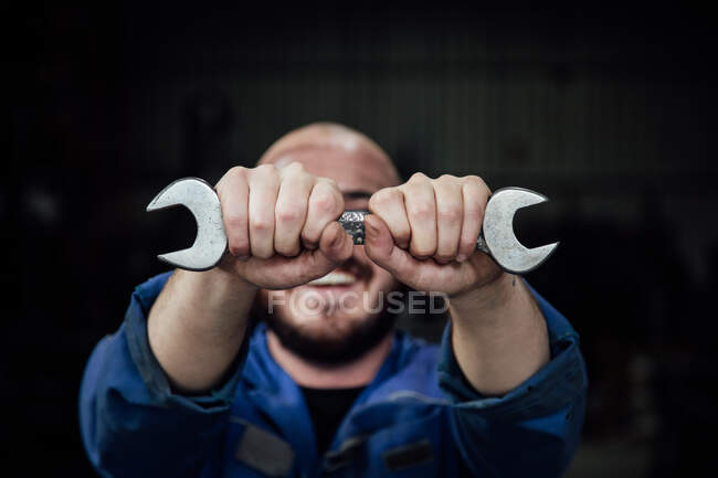Faceless male mechanic in blue overall covering face with metal spanner in hands looking at camera on black background — Stock Photo