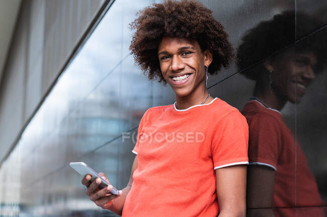 Content young African American male student with curly hair in t shirt messaging on mobile phone while standing near tiled wall on city street — Stock Photo