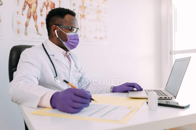 Médico afroamericano en anteojos que trabaja con paciente en línea en netbook mientras escribe en archivo de paciente en la mesa en el hospital - foto de stock
