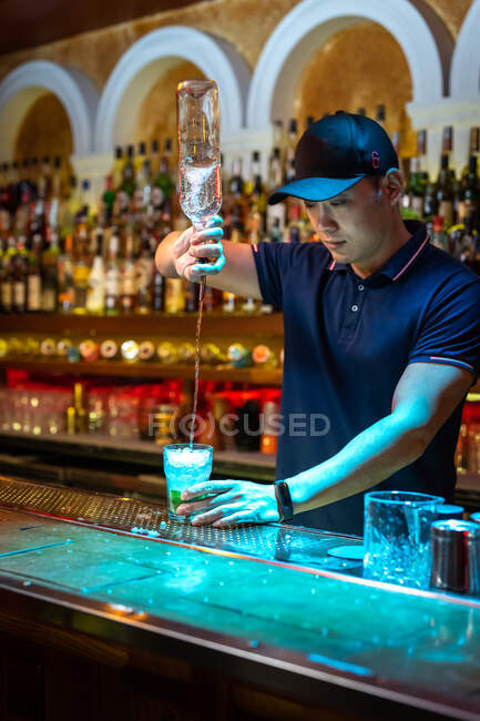 Joven camarero asiático vertiendo ron en el vaso mientras prepara cóctel mojito en el bar - foto de stock