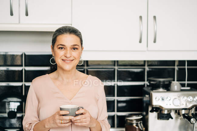 Весёлая женщина средних лет с кружкой горячего напитка, стоящей у кухонного стола с белыми шкафами и современной кофеваркой дома — стоковое фото