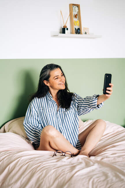 Полное тело веселой женщины среднего возраста в полосатой рубашке, улыбающейся и делающей селфи на смартфоне во время отдыха на кровати в выходные дни дома — стоковое фото