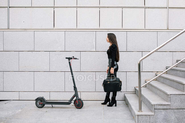 Vista laterale di fiduciosa imprenditrice alla moda in piedi vicino a scooter elettrico contemporaneo sulla strada della città — Foto stock