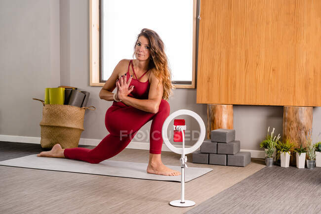 Ganzkörper-fitte Frau mit langen Haaren, die Revolved Crescent Lunge auf der Knie-Asana durchführt, während sie Yoga-Training auf dem Smartphone filmt, das an einem Stativ befestigt ist — Stockfoto