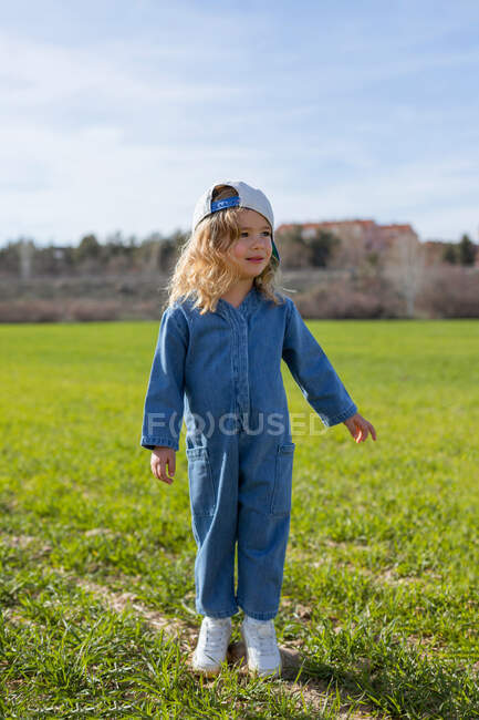 Ganzkörpermädchen in stylischer Kleidung und Mütze schaut weg, während sie an einem sonnigen Sommertag auf dem Feld auf dem Gras steht — Stockfoto