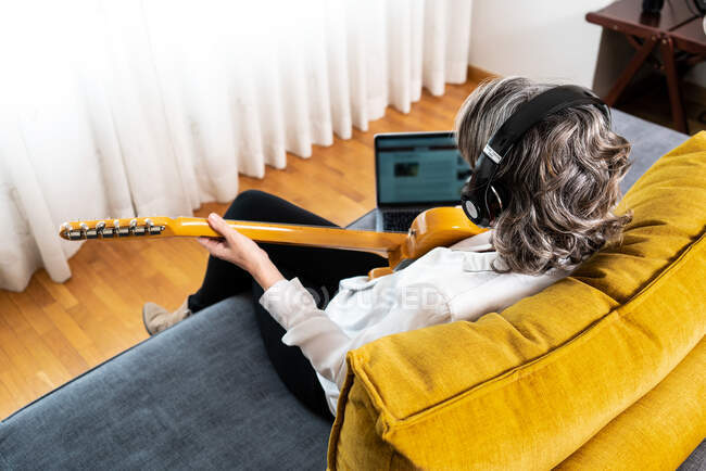 Vista trasera de ángulo alto de guitarrista femenina irreconocible en auriculares tocando instrumento musical en sofá con netbook en casa - foto de stock