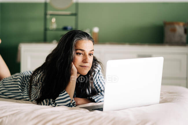 Вид сфокусированной зрелой самозанятой латиноамериканки с длинными темными волосами в повседневной одежде, лежащей на кровати с ноутбуком во время онлайн-работы дома — стоковое фото