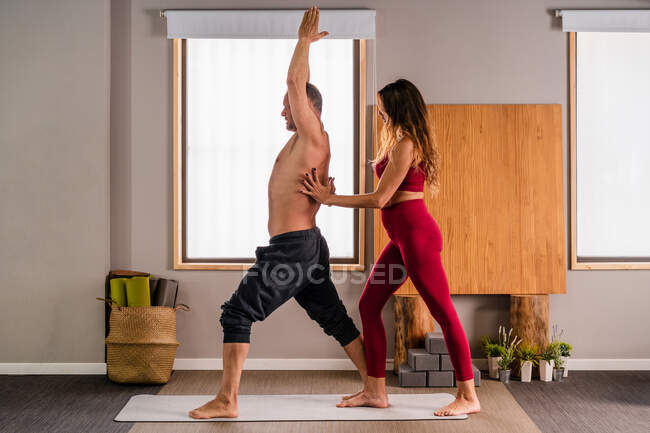 Vista lateral de la entrenadora femenina concentrada en ropa deportiva enseñando a hombre realizando pose de alta embestida durante la sesión de yoga en estudio - foto de stock