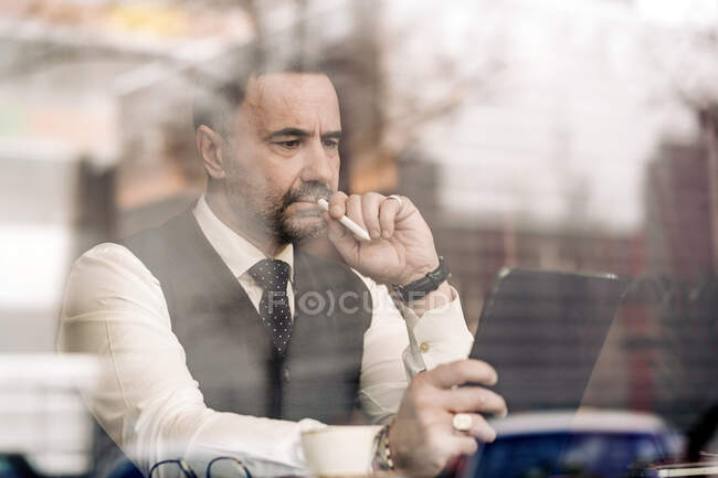 Продуманий етнічний підприємець середнього віку торкається рота зі стилусом, використовуючи планшет за скляною стіною в кафетерії — стокове фото