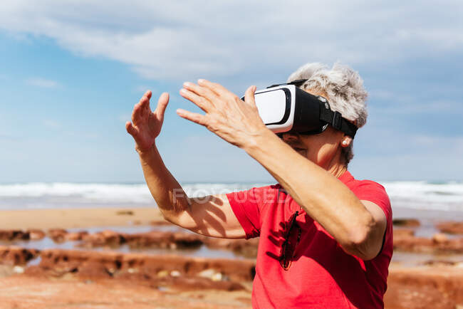 Anonyme ältere Reisende erleben virtuelle Realität mit Brille am Strand unter wolkenverhangenem Himmel — Stockfoto