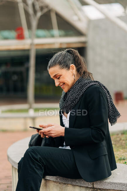 Side view of positive female text messaging on cellphone while sitting on stone border on street with constructions on blurred background — Stock Photo