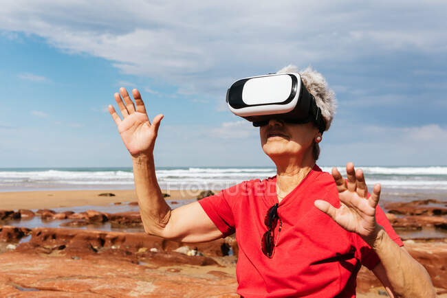 Anonymous elderly female traveler experiencing virtual reality in goggles on sea shore under cloudy sky — Stock Photo