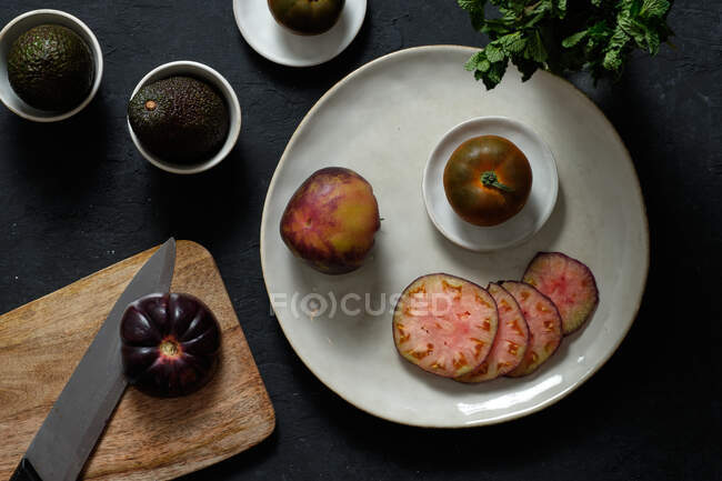 Top view of fresh whole and sliced black tomatoes on table with avocado and green mint during healthy meal preparation — Stock Photo