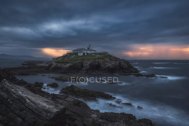 Breathtaking scenery of rocky island with lighthouse located in ocean near rocky coast in Faro Tapia de Casariego in Asturias in Spain under cloudy sky at sunrise — Stock Photo