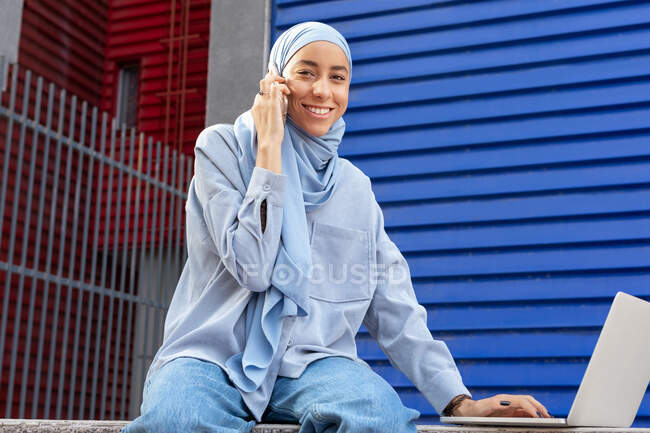 Mulher muçulmana sorridente em lenço de cabeça com netbook falando no celular enquanto olha para a câmera na cidade — Fotografia de Stock