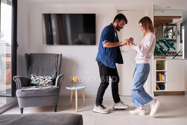 Vista lateral de la excitada pareja joven multirracial con ropa elegante cogida de la mano mientras bailan juntos en un apartamento moderno durante el día - foto de stock