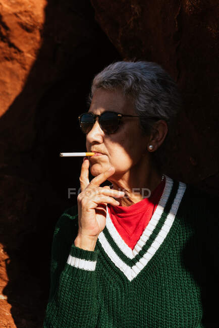 Elderly female with gray hair in knitted sweater with ornament and  sunglasses smoking cigarette — contemplative, warm clothes - Stock Photo |  #467857294