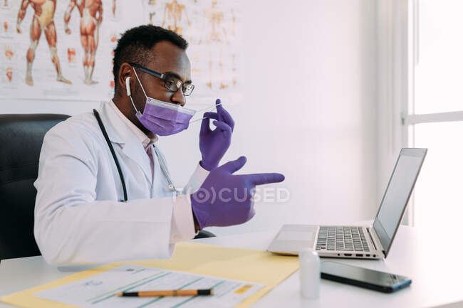 African American doctor in eyeglasses and face mask working with online patient on netbook while writing in patient file at table in hospital — Stock Photo