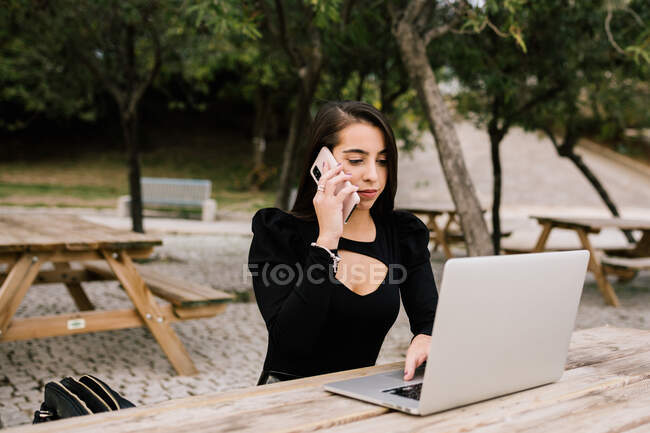 Female entrepreneur sitting at table with laptop in park and speaking on smartphone while working remotely — Stock Photo