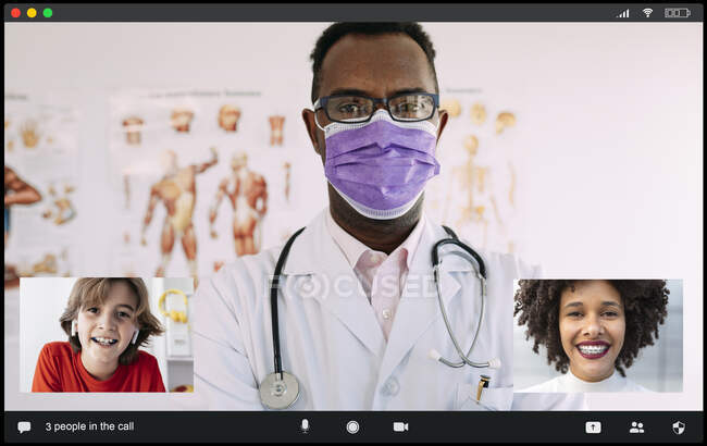 Нерозпізнані концентровані афроамериканський лікар чоловічої статі в медичному вбранні і маска дивлячись на камеру при спілкуванні з позитивною багатонаціональною жінкою і хлопчиком під час відео дзвінка — стокове фото