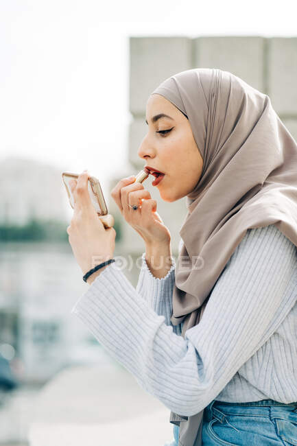 Seitenansicht einer Muslimin mit Kopftuch, die Lippenstift aufträgt und beim Schminken auf der Straße auf den Bildschirm ihres Smartphones schaut — Stockfoto