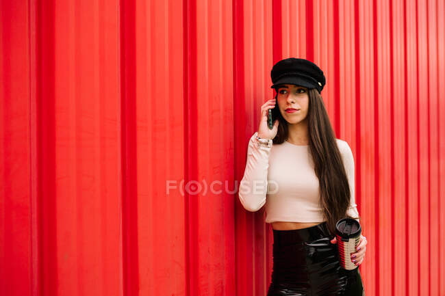 Serious stylish female entrepreneur leaning on red wall in city and discussing project on smartphone while standing with drink to go — Stock Photo