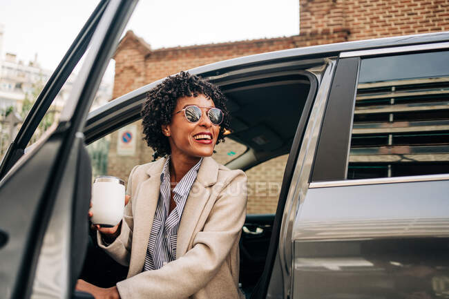 Mulher afro-americana feliz em óculos de sol na moda com caneca térmica sorrindo e deixando automóvel prestígio prata — Fotografia de Stock