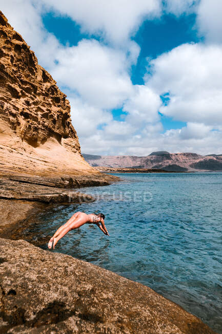 Corpo inteiro de viajante feminina em trajes de banho pulando em água ondulante do mar cercada por formações rochosas — Fotografia de Stock