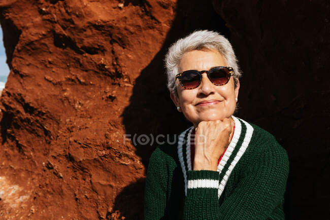 Happy elderly female tourist in casual apparel leaning on hand sitting on rocky formation with barren terrain under blue cloudy sky — Stock Photo