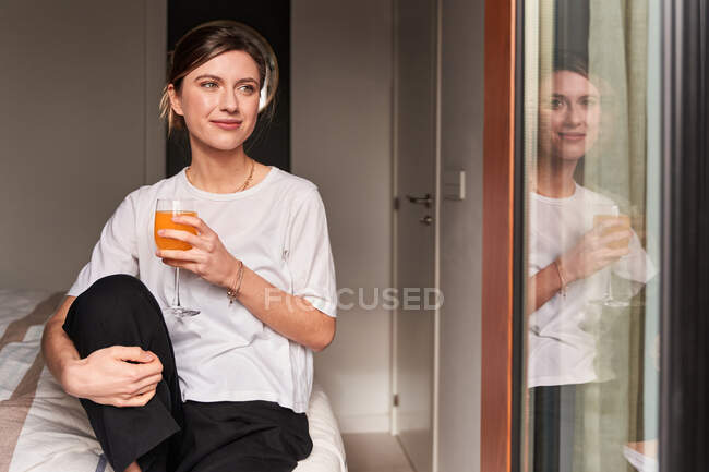 Content young female in casual clothes smiling and drinking fresh juice sitting on comfortable bed near window — Stock Photo