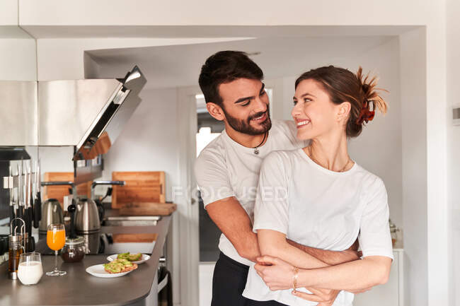 Вид сбоку молодого романтичного этнического парня в белой футболке, улыбающегося и обнимающего счастливую подругу, глядя друг на друга — стоковое фото