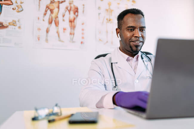 Jeune médecin afro-américain concentré en peignoir médical et écouteurs TWS travaillant sur ordinateur portable assis à table dans une clinique moderne — Photo de stock