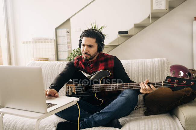 Взрослый музыкант в наушниках играет на бас-гитаре против нетбука на диване в гостиной — стоковое фото