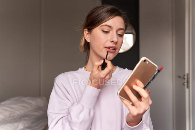 Щаслива молода жінка-блогер в повсякденному одязі малює губи під час зйомок відео на смартфоні для відеоблогу — стокове фото