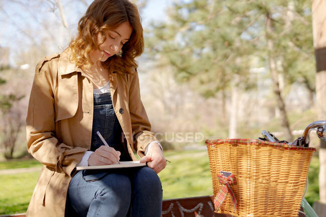 Joven mujer reflexiva tomando notas en planificador en banco de madera cerca de la bicicleta en el parque durante el día - foto de stock