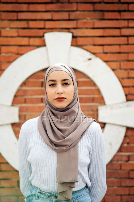 Femme musulmane charmante en foulard traditionnel et vêtements tendance debout sur la rue contre un mur de briques et regardant la caméra — Photo de stock