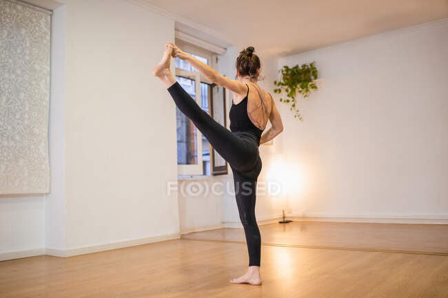 Side view of anonymous flexible barefoot female performing Extended Standing Hand to Toe pose on floor against luminous lamp — Stock Photo