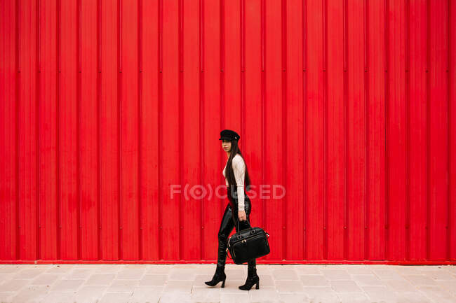 Вид збоку на жіночого підприємця в модному одязі, що йде вулицею на фоні червоної стіни і дивиться на камеру — стокове фото