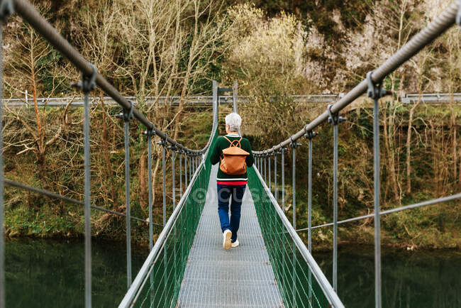 Back view of elderly female hiker in casual clothes strolling on suspension bridge in daytime — Stock Photo