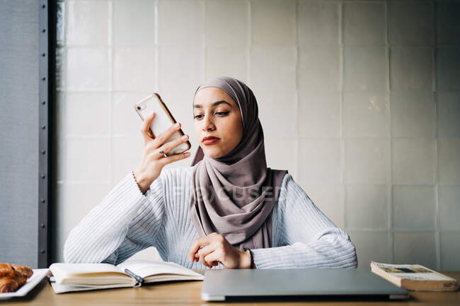 Contenido mujer freelancer étnica en hijab grabación de mensajes de audio en el teléfono inteligente mientras está sentado en la mesa en la cafetería y trabajando de forma remota - foto de stock