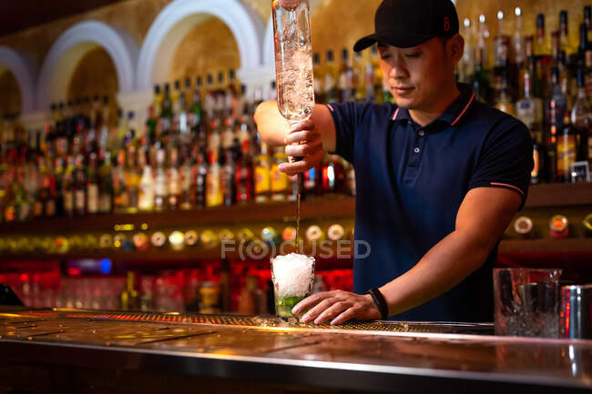Junger asiatischer Barkeeper gießt Rum ins Glas, während er in der Bar einen Mojito-Cocktail zubereitet — Stockfoto