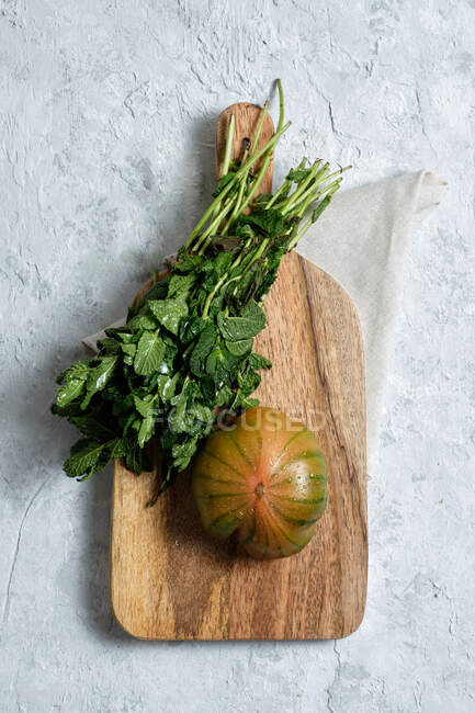 Desde arriba de tomate fresco rayado verde y rojo colocado en la tabla de cortar de madera con racimo de tallos de menta - foto de stock