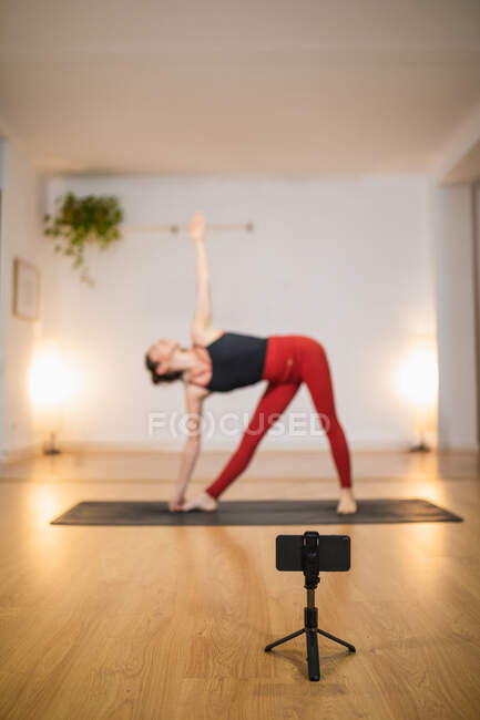 Vue latérale à angle élevé de la femelle mince à Trikonasana pose et tournage vidéo en direct sur smartphone pendant l'entraînement de yoga — Photo de stock