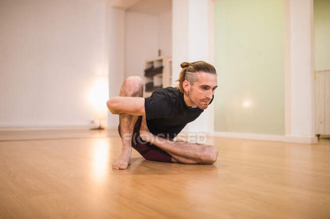 Adult male in sportswear with hands behind back performing forward bend while stretching legs on parquet — Stock Photo