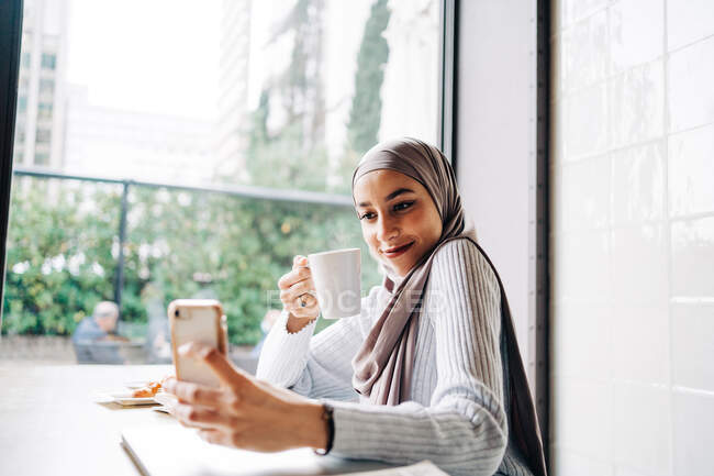 Щаслива етнічна жінка в хіджабі і з чашкою напою, приймаючи селфі на смартфон, насолоджуючись вихідними в кафе — стокове фото