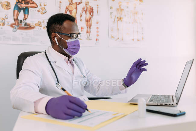 Médecin afro-américain dans les lunettes de travail avec le patient en ligne sur netbook tout en écrivant dans le dossier patient à la table à l'hôpital — Photo de stock