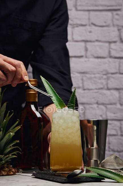 Crop unrecognizable bartender garnishing cocktail in glass with green leaves placed on slate coaster near shaker and bottle on counter against brick wall — Stock Photo